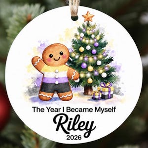 Könnte beinhalten: Ein runder, weißer Weihnachtsbaumschmuck mit einer Aquarellillustration eines Lebkuchenmanns im Muster der nicht-binären Flagge, einem geschmückten Weihnachtsbaum und zwei Geschenken. Der Text lautet The Year I Became Myself Riley 2026.
