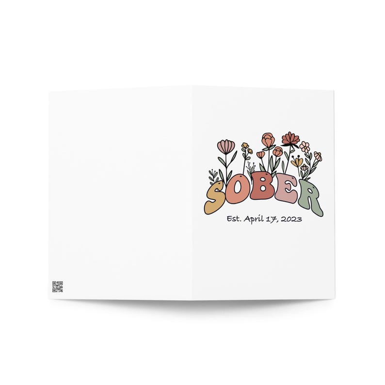 May include: A folded white greeting card with the word "SOBER" in colorful, retro-style lettering, accompanied by floral illustrations. The text "Est. April 17, 2023" is below the word "SOBER".