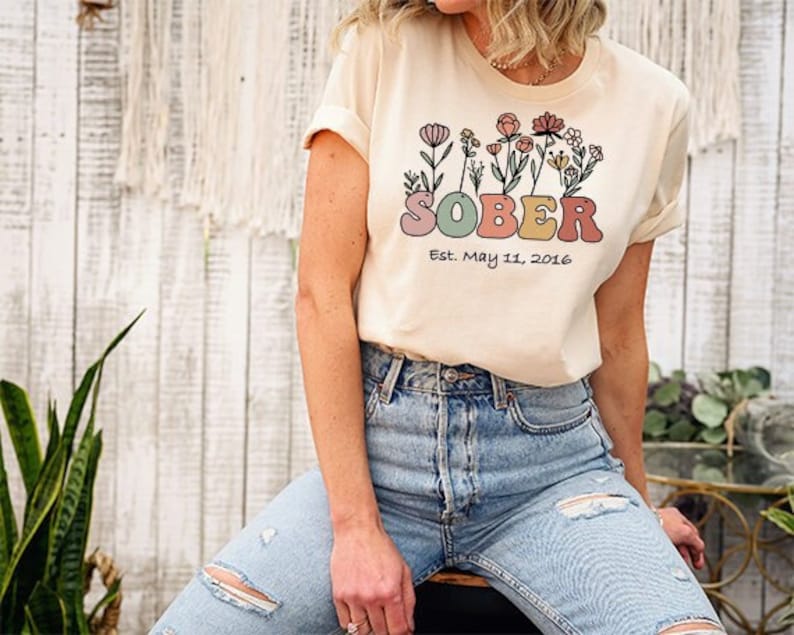 May include: Cream-colored t-shirt with the word "SOBER" in pastel colors, adorned with floral illustrations. The text "Est. May 11, 2016" is below the word "SOBER". The person is wearing blue jeans with ripped knees.