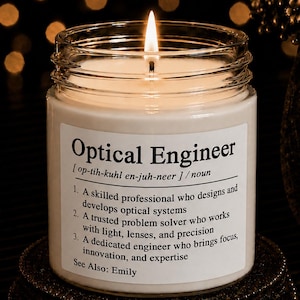 May include: A clear glass jar candle with a lit wick and white wax. The label features the text Optical Engineer, a phonetic pronunciation, the noun classification, and three definitions describing the profession, ending with See Also: Emily.