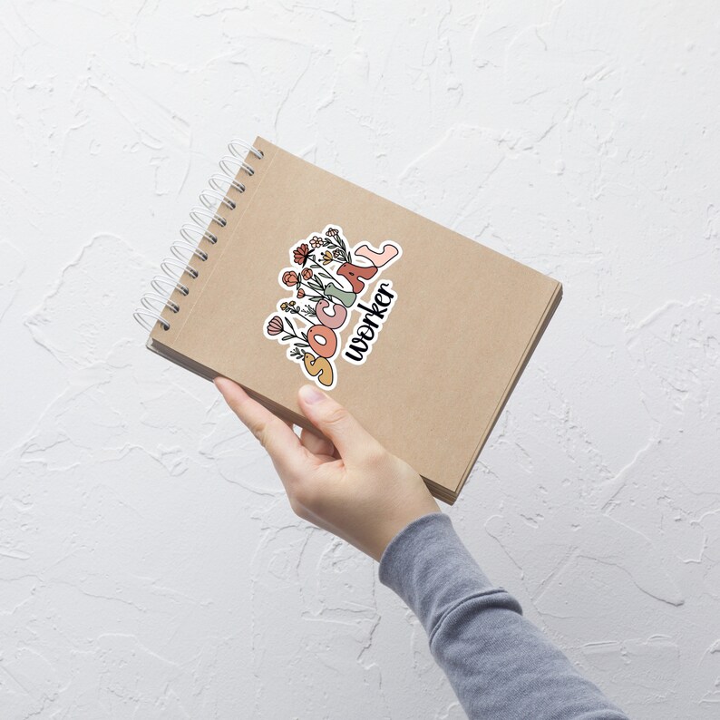 K&ouml;nnte beinhalten: Ein braunes Notizbuch mit Spiralbindung und einem Aufkleber, auf dem "SOCIAL worker" mit Blumen um den Text steht. Der Aufkleber ist in den Farben Rosa, Orange und Gr&uuml;n gehalten.