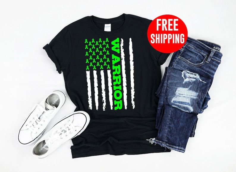 Op de afbeelding: Zwart T-shirt met een Amerikaans vlagontwerp met een groen lint en het woord "WARRIOR" verticaal gedrukt. Het shirt wordt getoond op een witte achtergrond met een paar witte sneakers en een blauwe jeans.