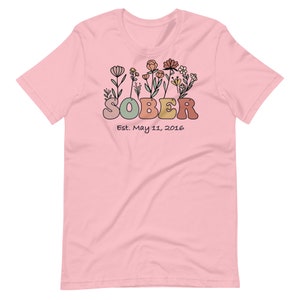 May include: A light pink t-shirt with a floral design above the word "SOBER" in retro-style lettering. Below the word "SOBER" is the text "Est. May 11, 2016". The floral design includes various flowers in shades of orange, yellow, and green.