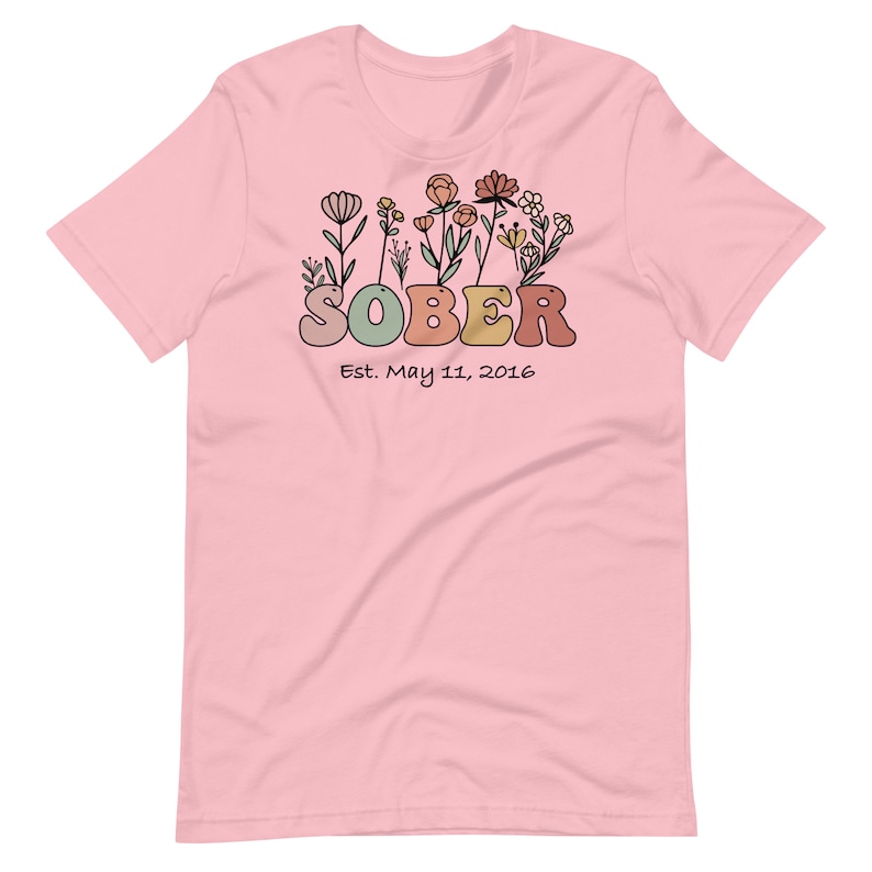 May include: A light pink t-shirt with a floral design above the word "SOBER" in retro-style lettering. Below the word "SOBER" is the text "Est. May 11, 2016". The floral design includes various flowers in shades of orange, yellow, and green.