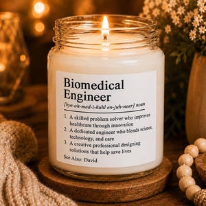 May include: A clear glass jar candle with a white wax fill and a lit wick. The label features the text Biomedical Engineer, a phonetic pronunciation, and three numbered definitions describing the profession, ending with the text See Also: David.