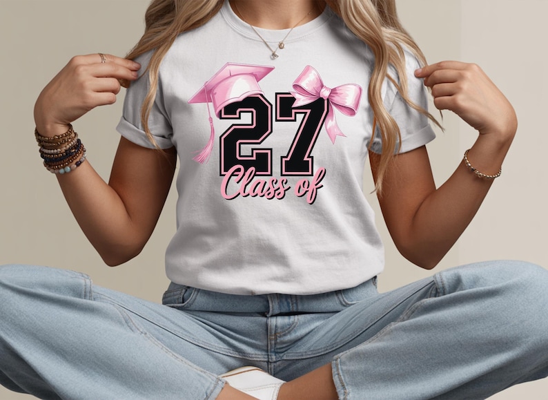 May include: White t-shirt featuring a pink graduation cap, a pink bow, and the number "27" in black with a pink outline. The words "Class of" are written in pink script below the number. The shirt is a graduation-themed apparel.