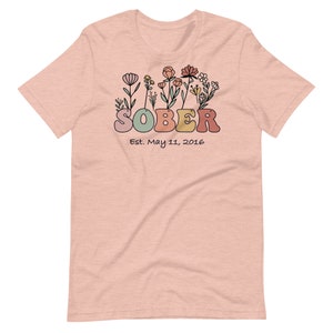 May include: A light pink t-shirt with a floral design and the word "SOBER" in colorful, retro-style letters. Below the word "SOBER" is the text "Est. May 11, 2016". The shirt is made of a soft material.