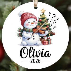 May include: A round white Christmas ornament featuring a snowman wearing a red knit hat and green scarf, playing a brown violin. Beside the snowman is a decorated Christmas tree with gifts. The name Olivia and the year 2026 are printed below.