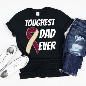 May include: Black t-shirt with the text "TOUGHEST DAD EVER" and a maroon and tan ribbon graphic. The shirt is paired with blue jeans and white sneakers.
