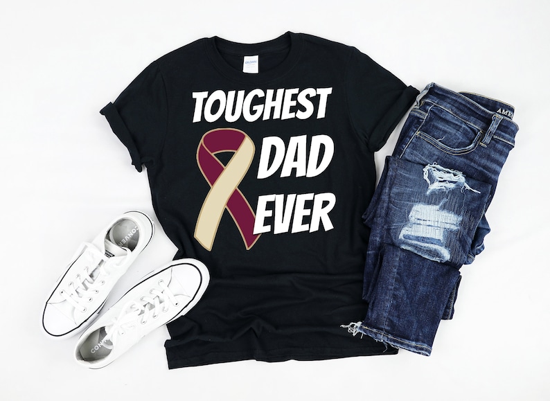 May include: Black t-shirt with the text "TOUGHEST DAD EVER" and a maroon and tan ribbon graphic. The shirt is paired with blue jeans and white sneakers.