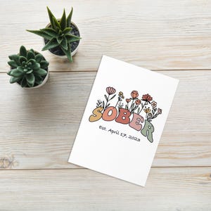 May include: A white card with the word "SOBER" in colorful bubble letters, surrounded by floral illustrations. Below the word is the text "Est. April 17, 2023". Two small potted succulents are in the background.