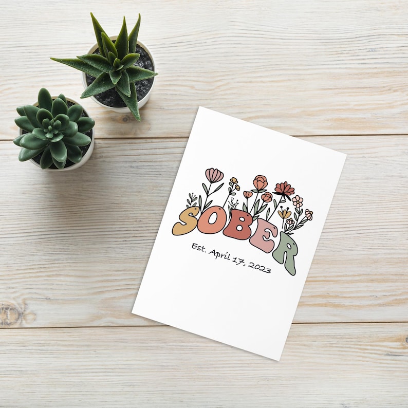 May include: A white card with the word "SOBER" in colorful bubble letters, surrounded by floral illustrations. Below the word is the text "Est. April 17, 2023". Two small potted succulents are in the background.