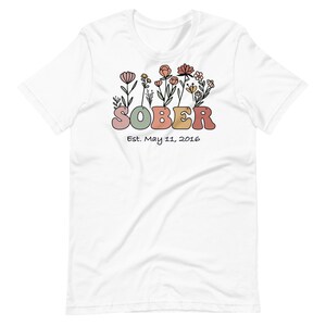 May include: White t-shirt with a floral design above the word "SOBER" in pastel colors. Below the word is the text "Est. May 11, 2016". The shirt has short sleeves and a crew neck.