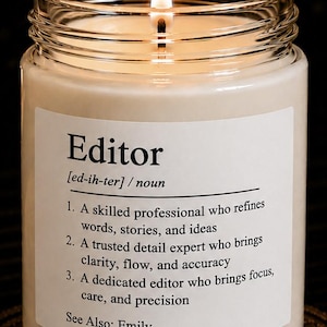 May include: A clear glass jar candle with a lit wick and a white label featuring the definition of the noun Editor. The text reads: Editor [ed-ih-ter] / noun 1. A skilled professional who refines words, stories, and ideas 2. A trusted detail expert who brings clarity, flow, and accuracy 3. A dedicated editor who brings focus, care, and precision. See Also: Emily.