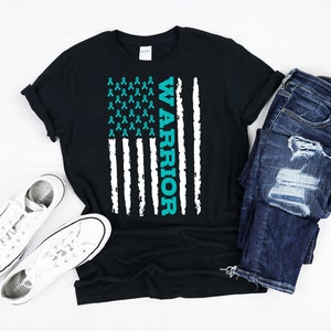 May include: A black t-shirt with a teal blue American flag design. The flag is made up of teal blue ribbon symbols and the word "WARRIOR" is written vertically on the right side of the flag.
