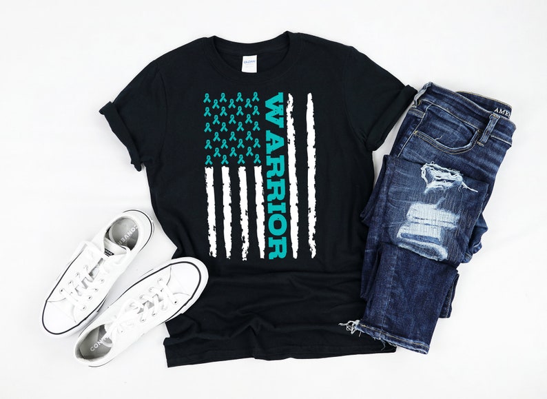 May include: A black t-shirt with a teal blue American flag design. The flag is made up of teal blue ribbon symbols and the word "WARRIOR" is written vertically on the right side of the flag.