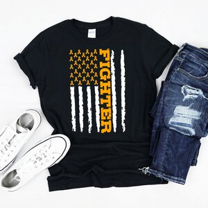 May include: Black t-shirt with an American flag design featuring orange ribbons and the word "Fighter". The shirt is displayed on a white surface with a pair of white sneakers and blue jeans.