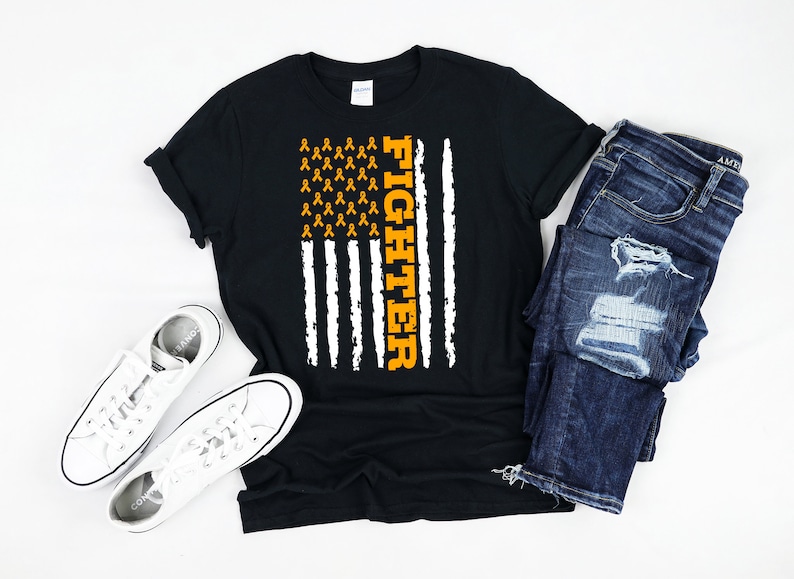May include: Black t-shirt with an American flag design featuring orange ribbons and the word "Fighter". The shirt is displayed on a white surface with a pair of white sneakers and blue jeans.
