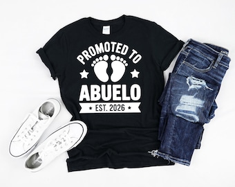 Puede incluir: Camiseta negra con el texto blanco "Promovido a Abuelo Est. 2026". El diseño incluye huellas de bebé y estrellas. La camiseta se combina con vaqueros azules y zapatillas blancas.