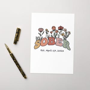May include: A white print featuring the word "SOBER" in colorful, rounded letters, with floral accents. Below the word is the text "Est. April 17, 2023". A gold and black pen and a pen are on the left side of the print.