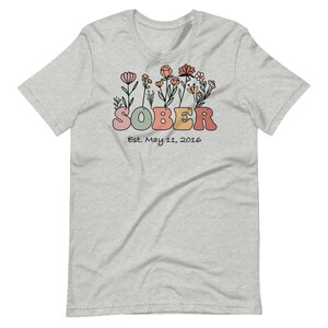 May include: Light gray t-shirt with a floral design above the word "SOBER" in pastel colors. The text "Est. May 11, 2016" is below the word "SOBER". The shirt is made of a soft material.