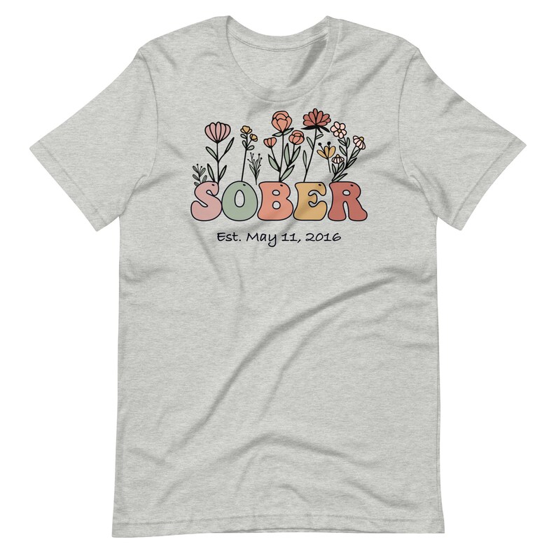 May include: Light gray t-shirt with a floral design above the word "SOBER" in pastel colors. The text "Est. May 11, 2016" is below the word "SOBER". The shirt is made of a soft material.
