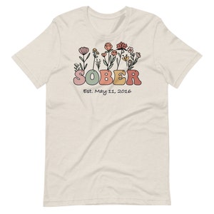 May include: A cream-colored t-shirt featuring the word "SOBER" in colorful, rounded letters, with a floral design above. Below the word is the text "Est. May 11, 2016". The shirt has short sleeves and a crew neck.