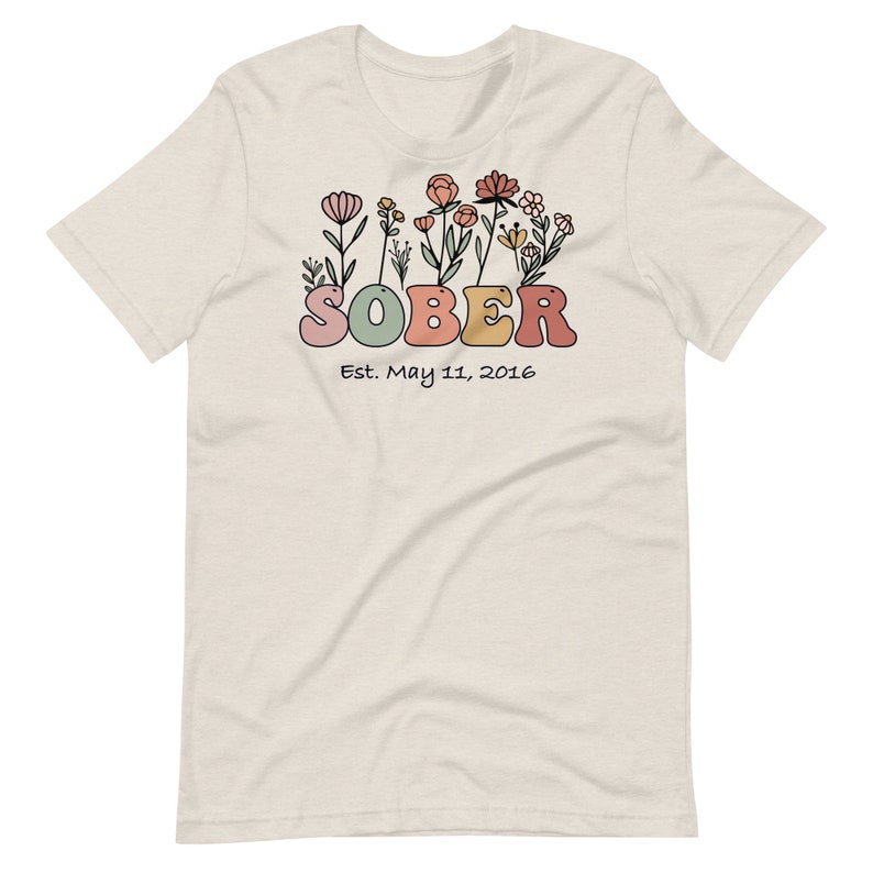 May include: A cream-colored t-shirt featuring the word "SOBER" in colorful, rounded letters, with a floral design above. Below the word is the text "Est. May 11, 2016". The shirt has short sleeves and a crew neck.