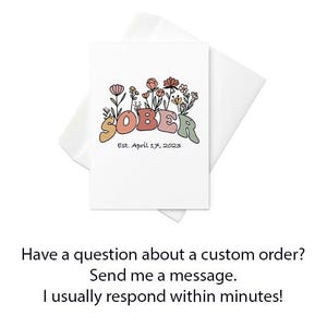 May include: A white greeting card with the word "SOBER" in retro-style lettering, adorned with floral illustrations in shades of orange, pink, and green. The card includes the text "Est. April 17, 2023". An envelope is behind the card.