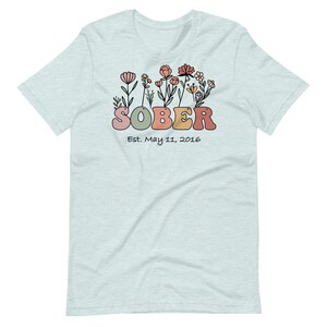May include: Light blue t-shirt with a floral design and the word "SOBER" in colorful letters. Below the word "SOBER" is the text "Est. May 11, 2016". The shirt is made of a soft material.