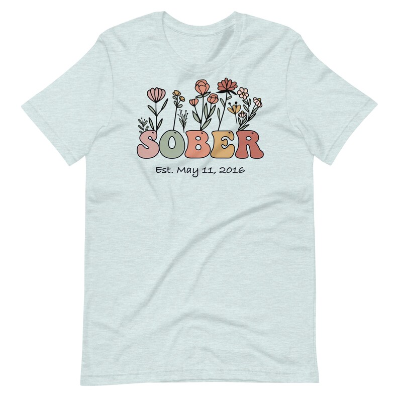 May include: Light blue t-shirt with a floral design and the word "SOBER" in colorful letters. Below the word "SOBER" is the text "Est. May 11, 2016". The shirt is made of a soft material.