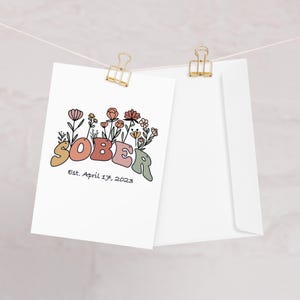 May include: A white card with the word "SOBER" in colorful, retro-style letters, adorned with floral illustrations. Below the word is the text "Est. April 17, 2023." The card is clipped to a string with a gold binder clip, next to a white envelope.