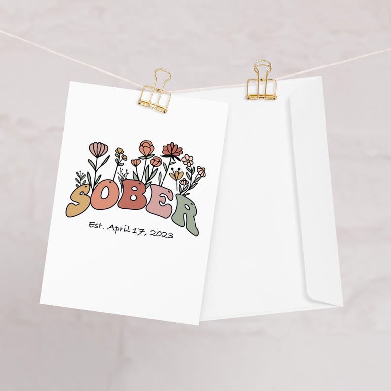 May include: A white card with the word "SOBER" in colorful, retro-style letters, adorned with floral illustrations. Below the word is the text "Est. April 17, 2023." The card is clipped to a string with a gold binder clip, next to a white envelope.