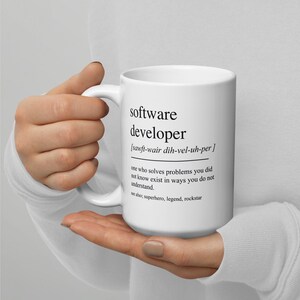 Op de afbeelding: Een witte keramische mok met een zwarte afbeelding die het woord "softwareontwikkelaar" definieert en een fonetische uitspraak bevat. De definitie luidt: "iemand die problemen oplost waarvan je niet wist dat ze bestonden, op een manier die je niet begrijpt." De tekst "zie ook: superheld, legende, rockstar" staat onder de definitie gedrukt.