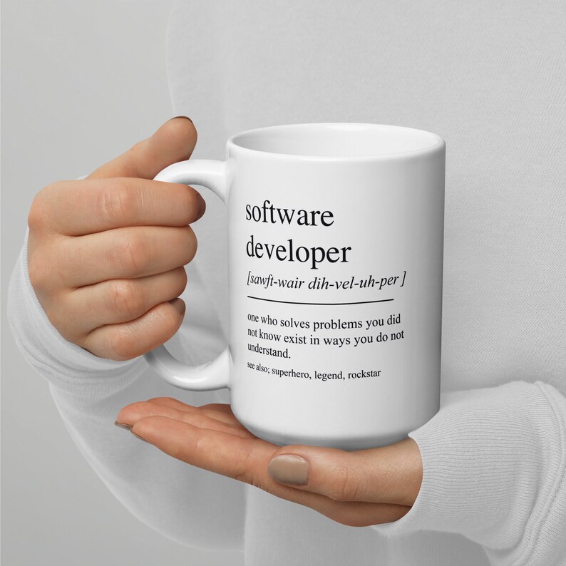 Op de afbeelding: Een witte keramische mok met een zwarte afbeelding die het woord "softwareontwikkelaar" definieert en een fonetische uitspraak bevat. De definitie luidt: "iemand die problemen oplost waarvan je niet wist dat ze bestonden, op een manier die je niet begrijpt." De tekst "zie ook: superheld, legende, rockstar" staat onder de definitie gedrukt.