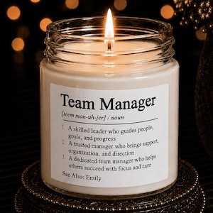 May include: A clear glass jar candle with a white wax fill and a lit wick. The label features the text Team Manager, a phonetic pronunciation, three numbered definitions describing a leader, and the text See Also: Emily in black font.