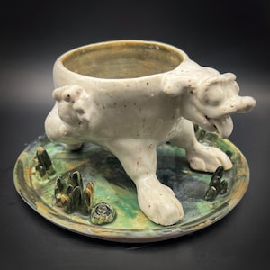 May include: A ceramic creature-shaped bowl with a glossy off-white glaze and speckled texture, featuring a protruding head with an open mouth and tongue. The bowl rests on a circular base decorated with green and blue swirls and small sculptures.