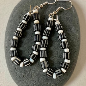 May include: A pair of teardrop-shaped earrings. Each earring is composed of alternating black and white beads. The black beads have white stripes. The earrings hang from silver-colored fish hook ear wires. The earrings are displayed on a gray stone.