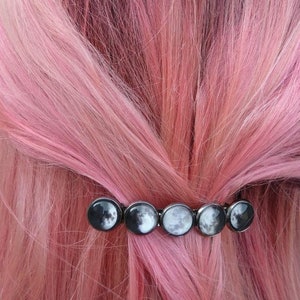 May include: A black hair clip with a row of silver moon phases. The clip is in pink hair.