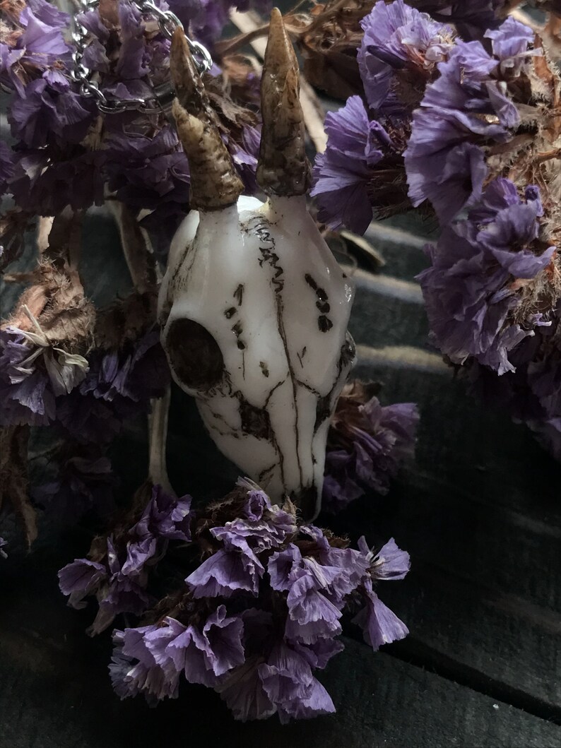 K&ouml;nnte beinhalten: Ein wei&szlig;er Keramik-Hirschsch&auml;del-Anh&auml;nger mit Geweih, der an einer silbernen Kette h&auml;ngt. Der Anh&auml;nger ist von getrockneten lila Blumen umgeben.
