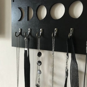 May include: A rectangular black wall-mounted organizer featuring five circular cutouts representing moon phases along the top edge. Five silver-toned metal hooks are attached to the lower section, each holding various necklaces and chokers.