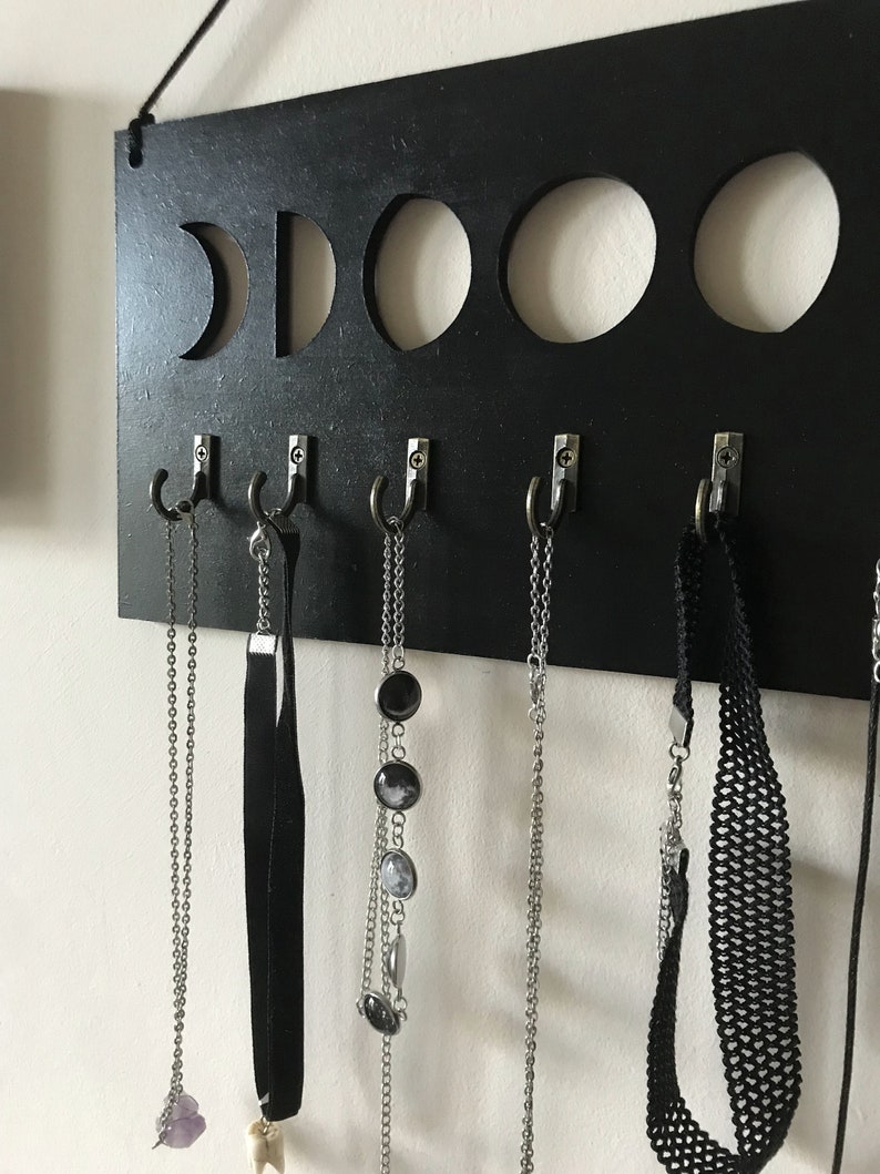 May include: A rectangular black wall-mounted organizer featuring five circular cutouts representing moon phases along the top edge. Five silver-toned metal hooks are attached to the lower section, each holding various necklaces and chokers.