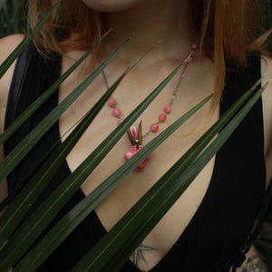 Pode incluir: Um colar de prata com contas rosa e um pingente com um design floral rosa e detalhes marrons. O colar &eacute; usado sobre um top preto. O logotipo Moonphases Jewellery &eacute; vis&iacute;vel.