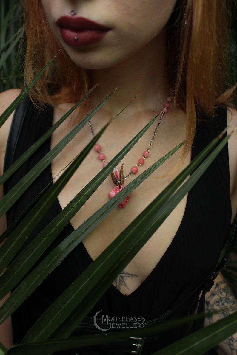 Pode incluir: Um colar de prata com contas rosa e um pingente com um design floral rosa e detalhes marrons. O colar &eacute; usado sobre um top preto. O logotipo Moonphases Jewellery &eacute; vis&iacute;vel.