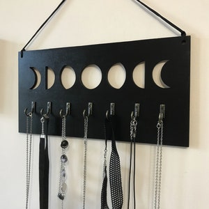 May include: A rectangular black wall-mounted jewelry organizer featuring a cutout moon phase design along the top edge. The board includes seven silver-toned metal hooks attached to the bottom, each holding various necklaces and chokers.