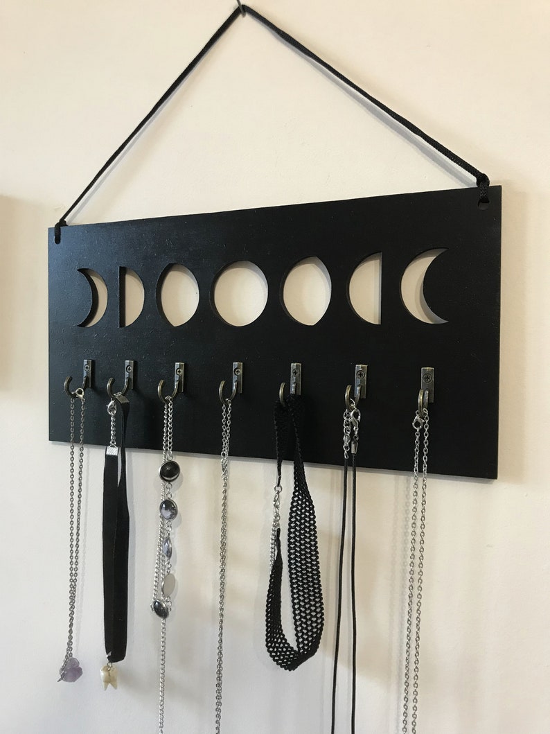 May include: A rectangular black wall-mounted jewelry organizer featuring a cutout moon phase design along the top edge. The board includes seven silver-toned metal hooks attached to the bottom, each holding various necklaces and chokers.