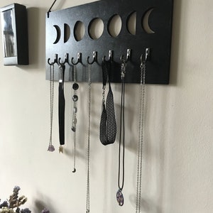 May include: A rectangular black wooden wall-mounted jewelry organizer featuring a cutout moon phase design along the top edge. The board includes seven silver-toned metal hooks, each holding a variety of necklaces, chains, and chokers.