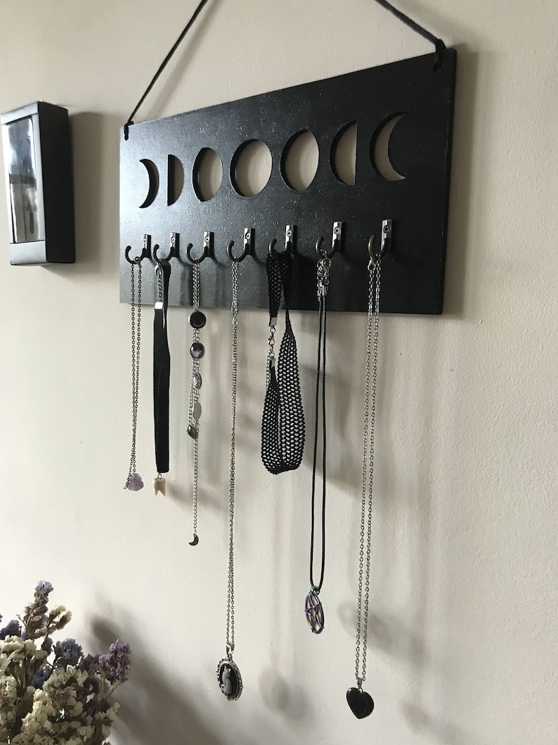 May include: A rectangular black wooden wall-mounted jewelry organizer featuring a cutout moon phase design along the top edge. The board includes seven silver-toned metal hooks, each holding a variety of necklaces, chains, and chokers.