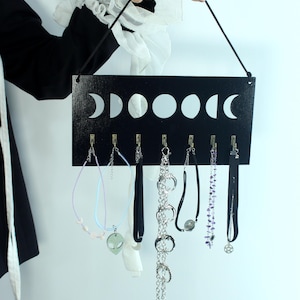 May include: A rectangular black wooden wall-mounted jewelry organizer featuring a white moon phase graphic across the top. It includes seven small metal hooks for hanging necklaces and chokers. The text "MOONPHASES JEWELLERY" is printed at the bottom.