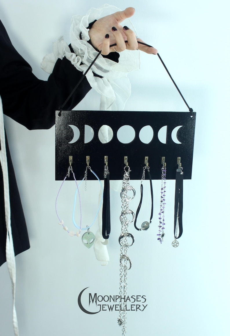 May include: A rectangular black wooden wall-mounted jewelry organizer featuring a white moon phase graphic across the top. It includes seven small metal hooks for hanging necklaces and chokers. The text "MOONPHASES JEWELLERY" is printed at the bottom.